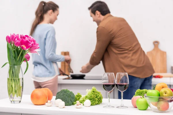 Various vegetables and fruits on table at kitchen with blurred couple cooking dinner together on background — Stock Photo