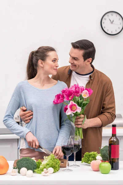 Bel homme adulte présentant bouquet de tulipes pour femme pendant qu'elle cuisine — Photo de stock