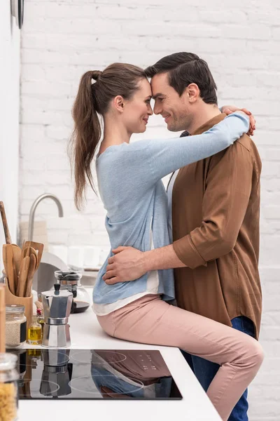 Feliz casal apaixonado abraçando na cozinha enquanto namorada sentada na mesa — Stock Photo