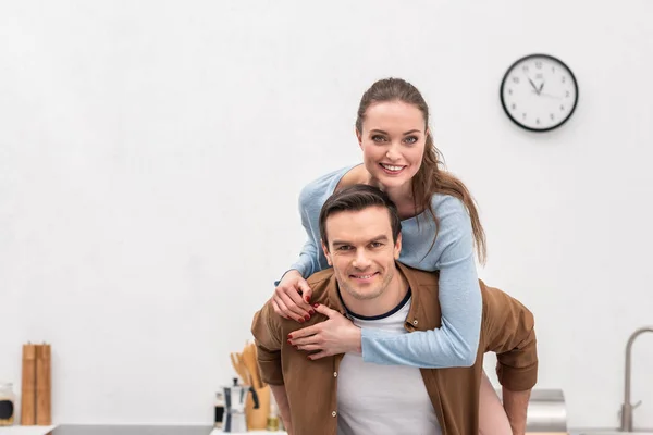 Feliz esposa piggybacking en marido en casa y mirando la cámara - foto de stock