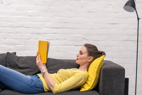 Seitenansicht einer erwachsenen Frau, die Buch liest, während sie zu Hause auf der Couch liegt — Stockfoto