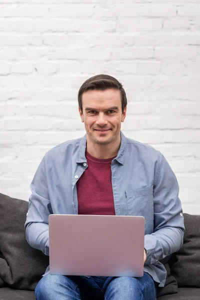 Homme adulte heureux en utilisant un ordinateur portable sur le canapé à la maison et en regardant la caméra — Photo de stock