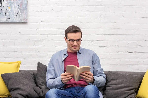 Happy adult man reading book on sofa at home in front of white brick wall — Stock Photo