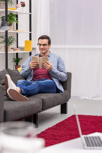 Hombre adulto sonriente leyendo libro en el sofá en casa - foto de stock