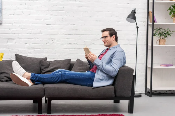 Visão lateral do homem adulto feliz livro de leitura no sofá em casa — Fotografia de Stock