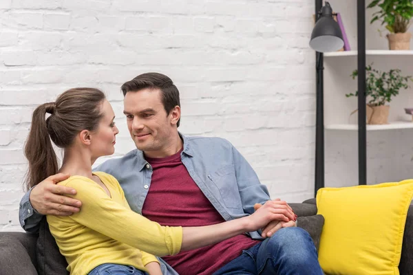 Feliz pareja adulta cogida de la mano mientras está sentada en el sofá en casa - foto de stock