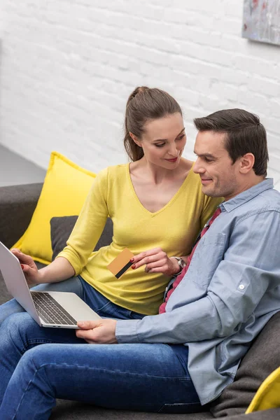 Sonriente pareja adulta haciendo e-shopping con laptop y tarjeta de crédito en el sofá en casa - foto de stock