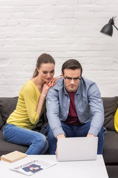 Attractive adult couple leaning on her husband while her working with laptop on couch at home — Stock Photo