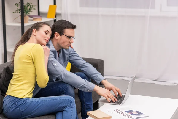 Beautiful adult couple leaning on her husband while her working with laptop on couch at home — Stock Photo