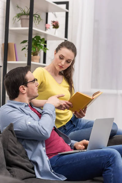 Привлекательная женщина с книгой в руках разговаривает с парнем в то время как он работает с ноутбуком на диване дома — стоковое фото