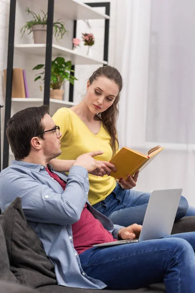 Donna attraente con libro in mano che parla con il fidanzato mentre lavora con il computer portatile sul divano a casa — Foto stock