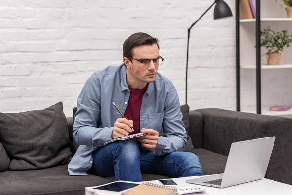 Fokussierter erwachsener Mann schreibt Notizen und schaut auf Laptop — Stockfoto