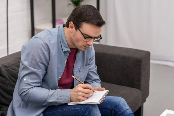 Concentrated adult man writing in notebook  while sitting on couch — Stock Photo