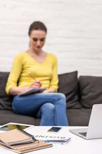 Gadgets sur la table avec une femme adulte floue travaillant avec un ordinateur portable à la maison sur le canapé sur le fond — Photo de stock