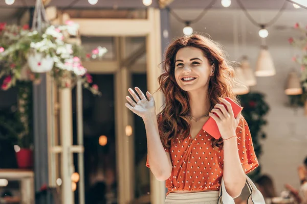 Portrait of cheerful woman with smartphone in hand waiving to someone on street — Stock Photo