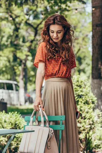 Портрет молодой женщины в стильной одежде кладет сумку на стул на улице — стоковое фото