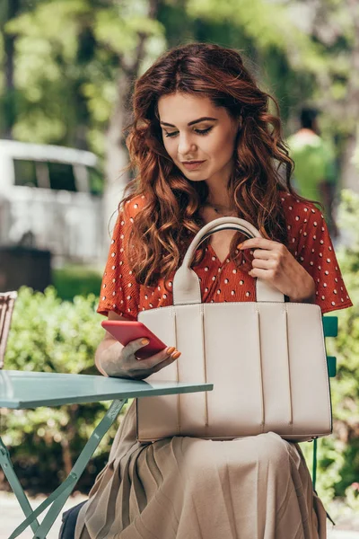 Портрет стильной женщины с сумкой на смартфоне, сидящей на террасе в кафе — стоковое фото