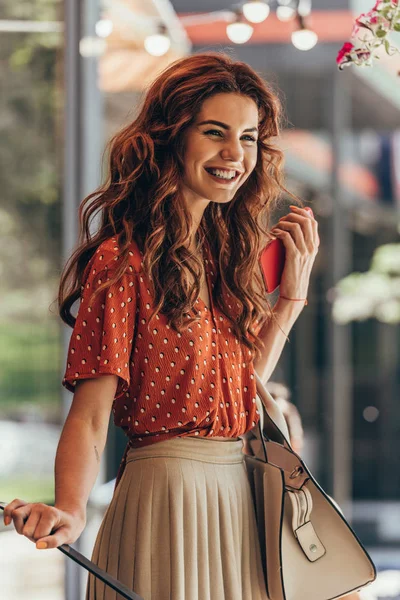 Vista lateral de la mujer sonriente en ropa elegante con teléfono inteligente y bolsa - foto de stock