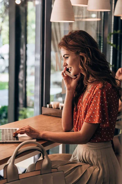 Vue latérale de la jeune femme joyeuse parlant sur smartphone à table avec ordinateur portable dans un café — Photo de stock