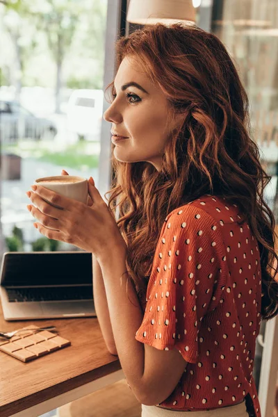 Vue latérale de femme coûteuse avec tasse de café à la table avec ordinateur portable dans le café — Photo de stock