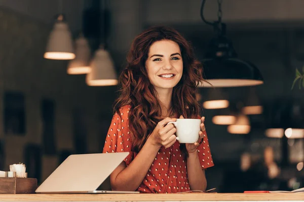 Portrait de femme souriante avec tasse de café à table avec ordinateur portable dans un café — Photo de stock