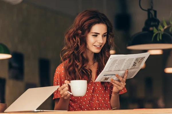 Ritratto di donna con tazza di caffè lettura giornale a tavola con computer portatile in caffè — Foto stock