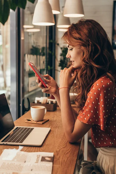 Vista lateral da jovem mulher usando smartphone à mesa com laptop no café — Fotografia de Stock