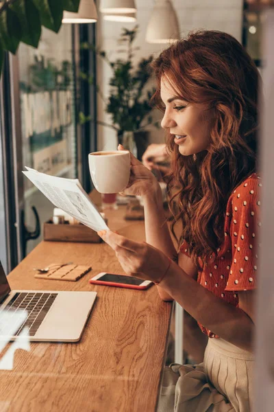 Vista lateral de la mujer con taza de café periódico de lectura en la mesa con el ordenador portátil en la cafetería - foto de stock