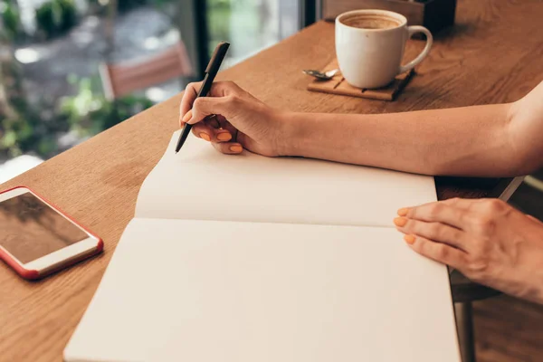 Обрезанный снимок блогера, делающего заметки в блокноте за столом с чашкой кофе в кафе — стоковое фото