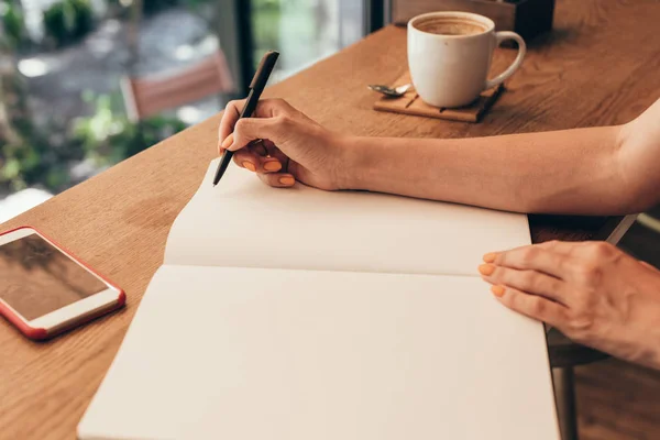 Recortado tiro de bloguero haciendo notas en el cuaderno en la mesa con taza de café en la cafetería - foto de stock