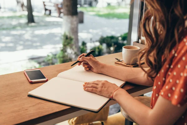 Plan recadré de blogueur prenant des notes dans un carnet à table avec une tasse de café dans un café — Photo de stock