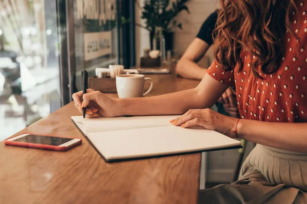 Partial view of blogger making notes in notebook at table with cup of coffee in coffee shop — Stock Photo