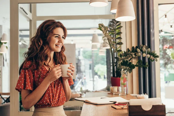 Улыбающаяся молодая женщина держит чашку кофе за столом с блокнотом в кафе — стоковое фото