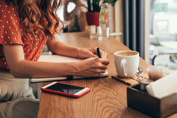 Recortado tiro de bloguero haciendo notas en el cuaderno en la mesa con taza de café en la cafetería — Stock Photo