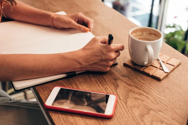 Partial view of blogger making notes in notebook at table with cup of coffee in coffee shop — Stock Photo