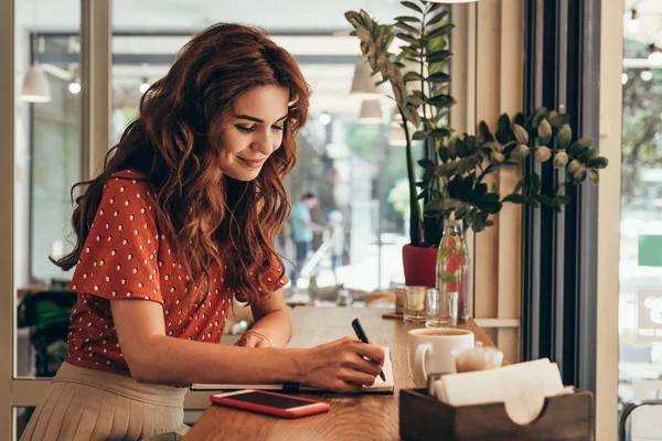 Vista laterale del giovane blogger che prende appunti nel quaderno a tavola con tazza di caffè in caffetteria — Foto stock