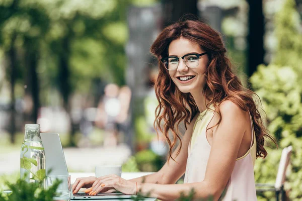Вид сбоку улыбающегося молодого блогера, смотрящего на камеру во время работы на ноутбуке в кафе — стоковое фото