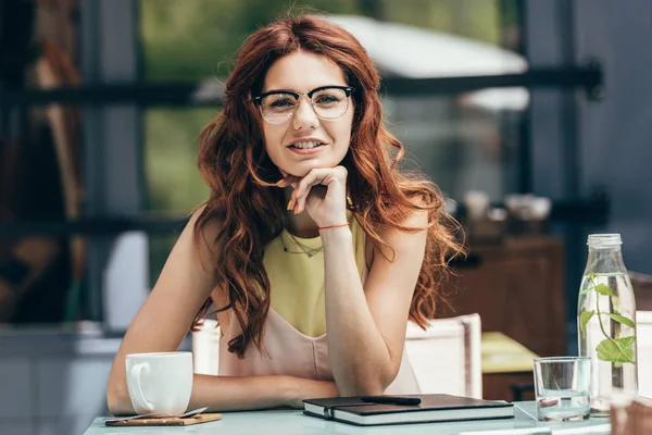 Retrato de atractiva mujer de negocios en anteojos sentado a la mesa con taza de café y portátil en la cafetería - foto de stock