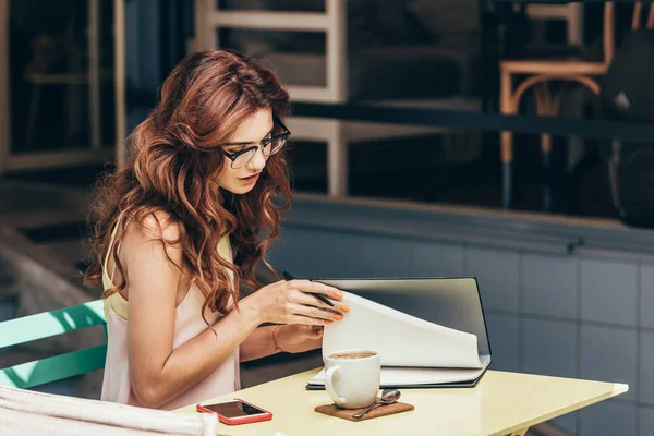 Vista lateral de la joven mujer de negocios en gafas con cuaderno en la mesa en la cafetería - foto de stock