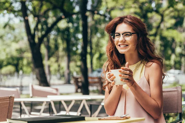 Empresária alegre em óculos com xícara de café sentado à mesa com notebook no café — Fotografia de Stock