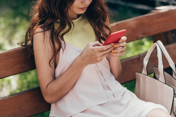 Vista parcial de la mujer con estilo utilizando el teléfono inteligente mientras descansa en el banco - foto de stock