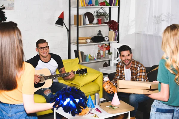 Giovani uomini sorridenti che suonano la chitarra e guardando le ragazze che tengono la pizza a casa festa — Foto stock