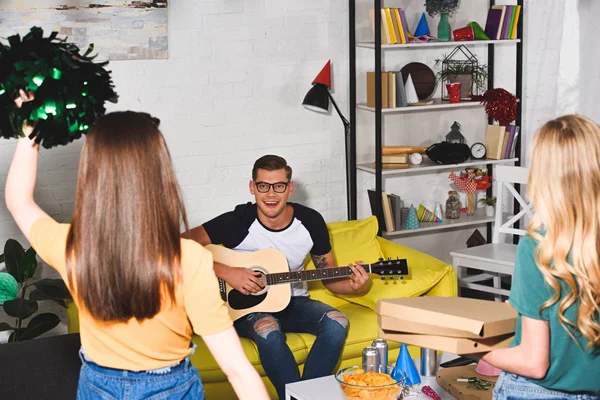 Visão traseira de meninas com caixas de pizza e pom-poms olhando para o homem sorridente tocando guitarra na festa em casa — Fotografia de Stock