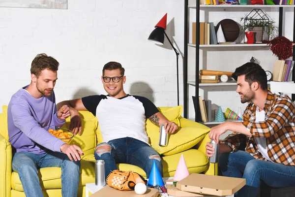 Gutaussehende lächelnde junge Männer, die Bier trinken und zu Hause Snacks essen — Stockfoto