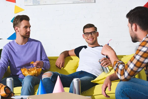 Улыбающиеся молодые мужчины пьют пиво и едят закуски на домашней вечеринке — стоковое фото