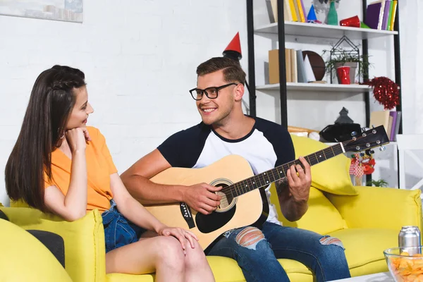 Guapo sonriente joven tocando la guitarra acústica y mirando a la hermosa chica sentada en el sofá - foto de stock
