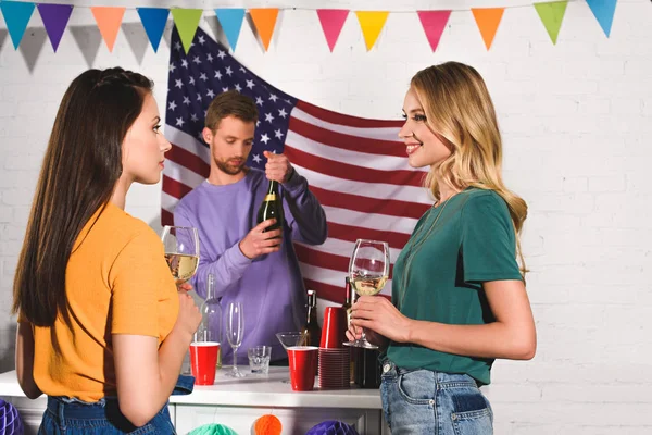 Beautiful young women holding glasses of wine and looking at each other while man opening wine bottle behind — Stock Photo