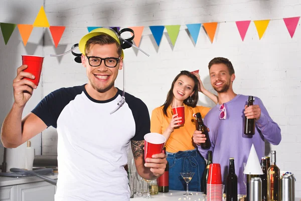 Beau jeune homme en chapeau de bière tenant des tasses en plastique et souriant à la caméra tout en faisant la fête avec des amis — Photo de stock