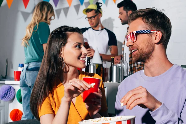 Счастливый молодой человек и женщина улыбаются друг другу, когда пьют пиво и едят попкорн на домашней вечеринке — стоковое фото
