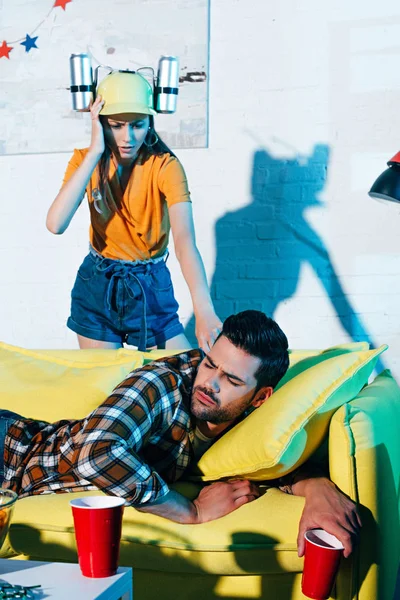 Shocked girl in beer hat looking at drunk man lying on sofa after home party — Stock Photo